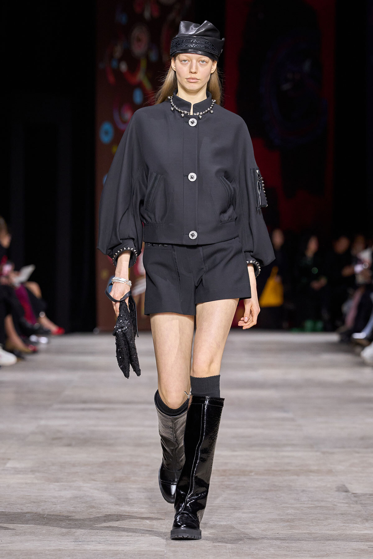 Model walks the runway in a dark gray oversized jacket with puff sleeves, matching shorts, black knee-high patent boots, and a black cap with pearl necklace.