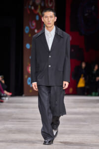 Male model walks a fashion runway in a black suit with a white shirt, silver-studded collar detail, and draped pants.