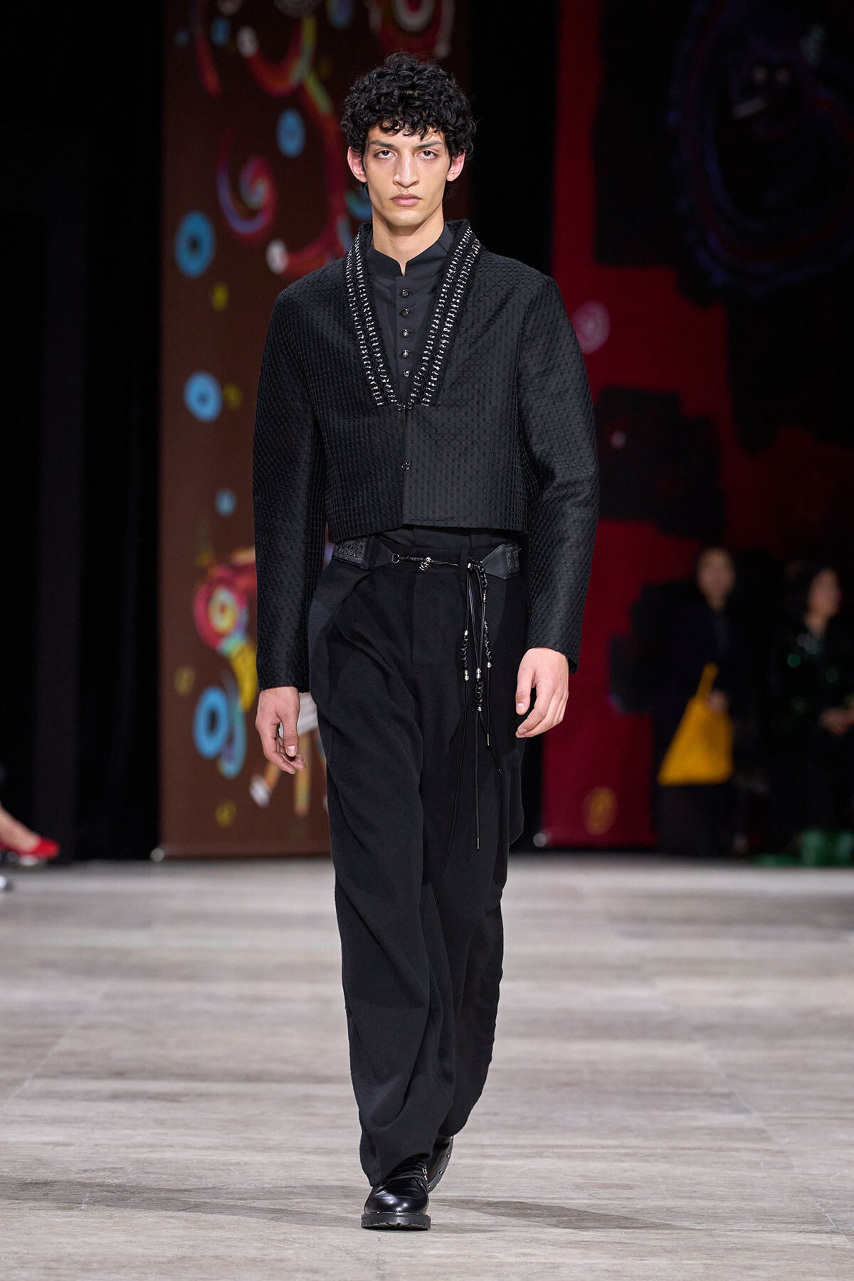 Male model on a runway wearing a textured black blazer with a V-trimmed neckline, black shirt, and loose black trousers with black loafers.