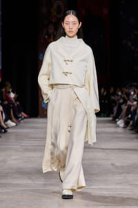 Model walks a fashion runway in an all-cream, layered ensemble featuring a cropped jacket with toggle closures and wide, flowing trousers.