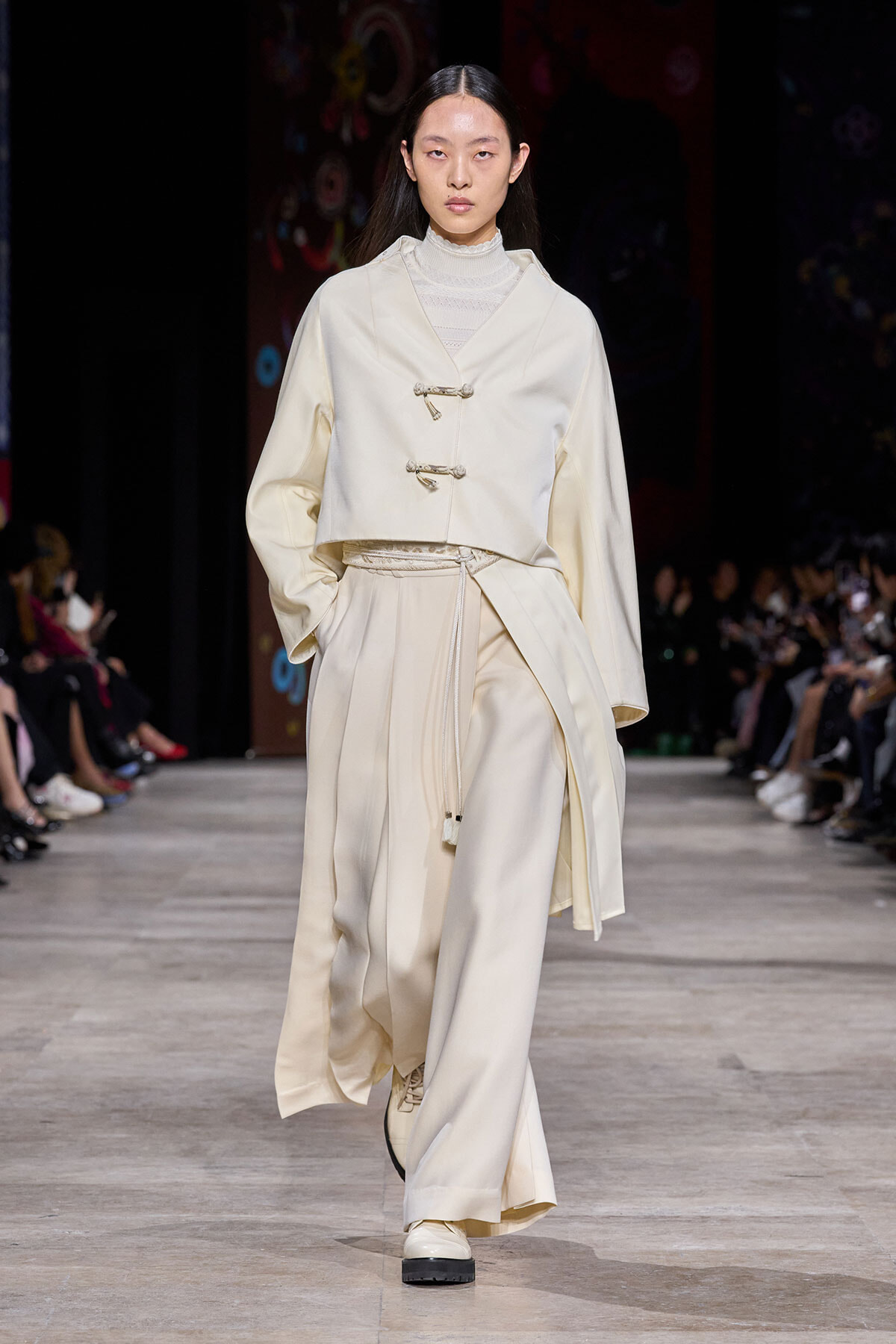 Model walks a fashion runway in an all-cream, layered ensemble featuring a cropped jacket with toggle closures and wide, flowing trousers.