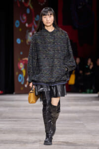 Model walks the runway in an oversized dark speckled knit sweater, black leather mini skirt, tall black boots, and a small tan handbag.