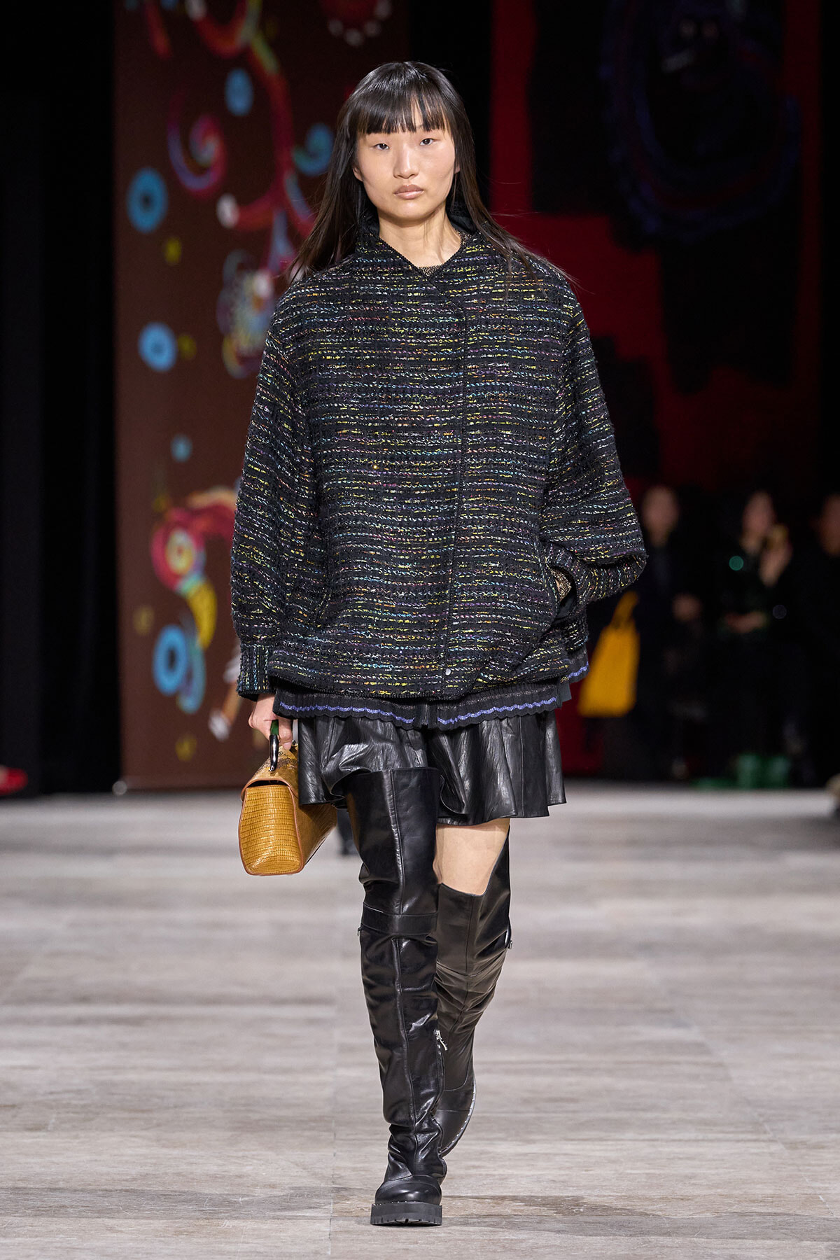 Model walks the runway in an oversized dark speckled knit sweater, black leather mini skirt, tall black boots, and a small tan handbag.