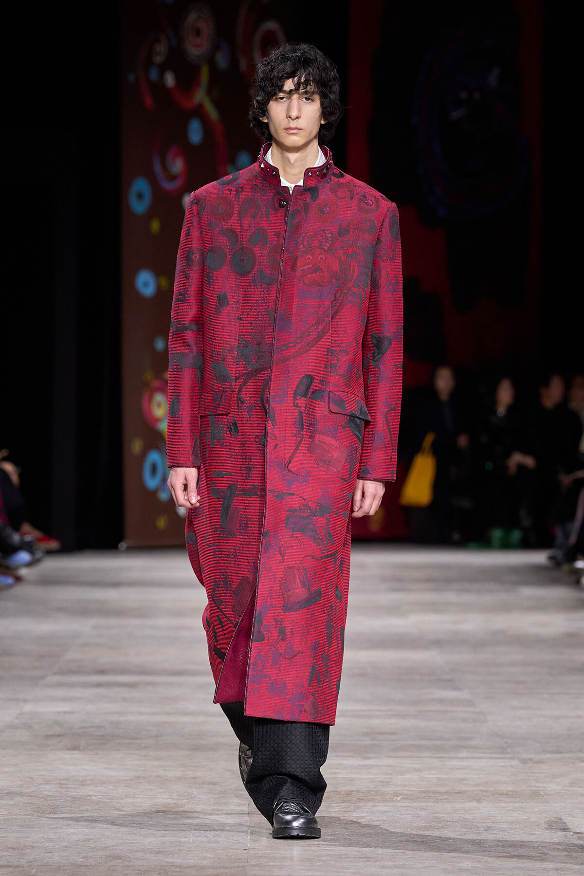 Male model walks a runway wearing a long red patterned coat over dark trousers and black shoes.
