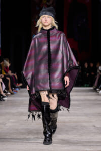 Model walking a fashion show runway in a metallic purple cape with black fur trim, paired with a black beanie and tall boots.