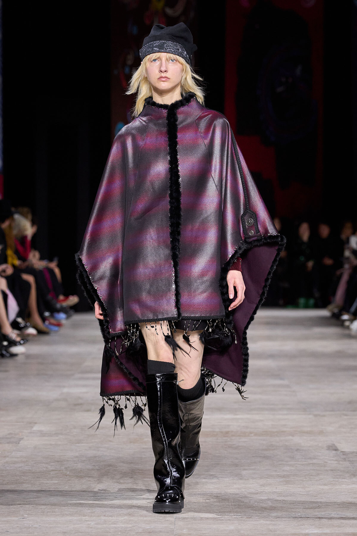 Model walking a fashion show runway in a metallic purple cape with black fur trim, paired with a black beanie and tall boots.
