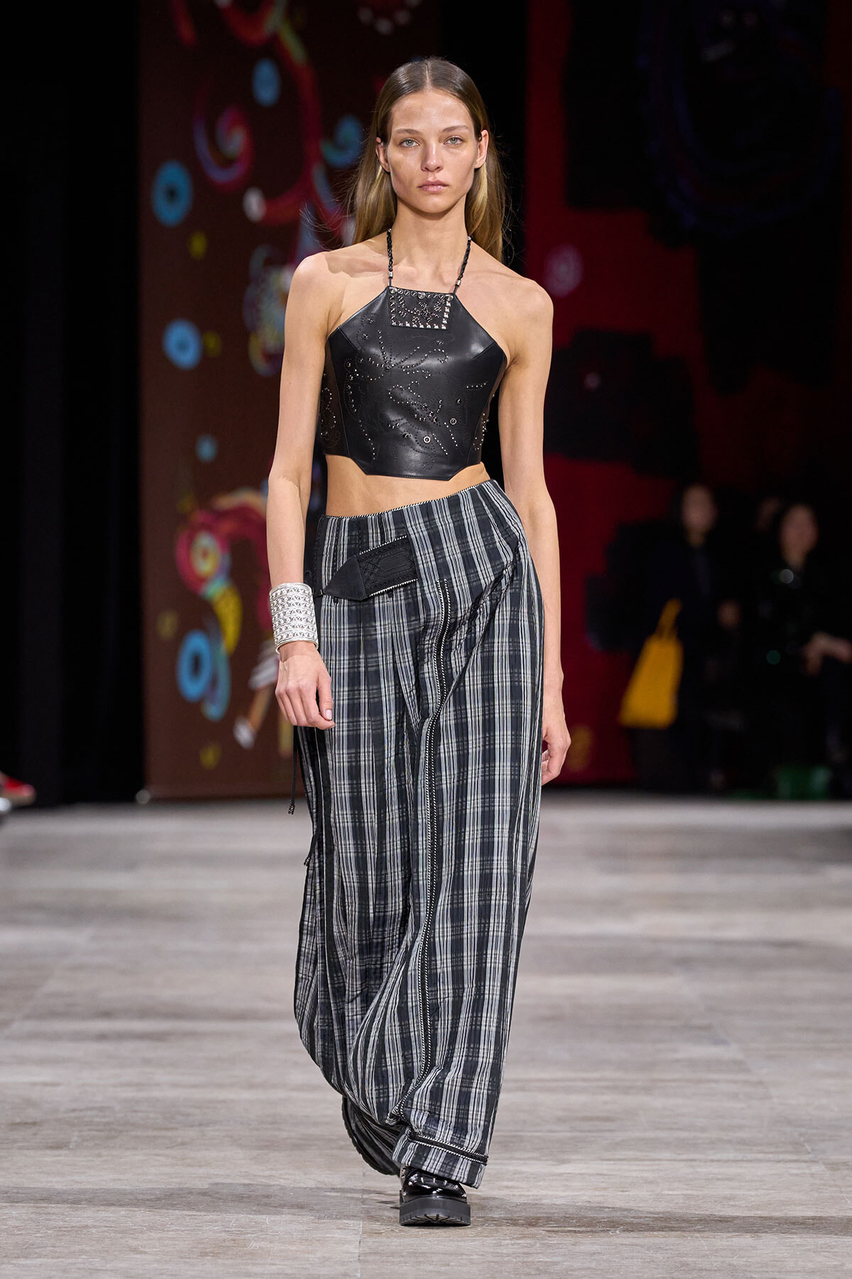 Model walks runway in a black leather halter crop top with studded neckline and loose plaid wide-leg pants.