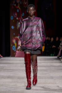 Model on a fashion runway wearing a burgundy-silver satin embroidered jacket, black shorts, tall red boots, and a pink clutch.