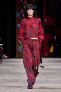 Model walks a runway in a burgundy lace blouse and patterned matching pants with decorative tassels and fringed belt.