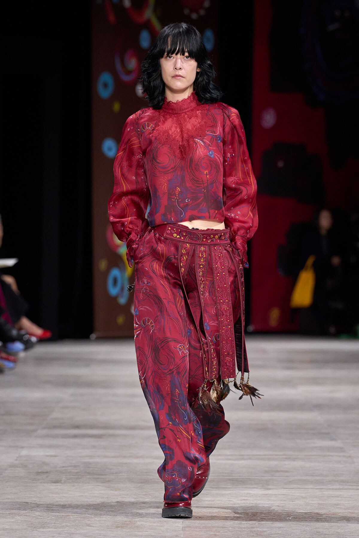 Model walks a runway in a burgundy lace blouse and patterned matching pants with decorative tassels and fringed belt.