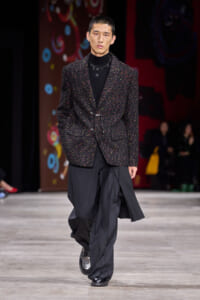 Male model walks a runway in a dark speckled blazer over a black turtleneck and oversized black trousers.