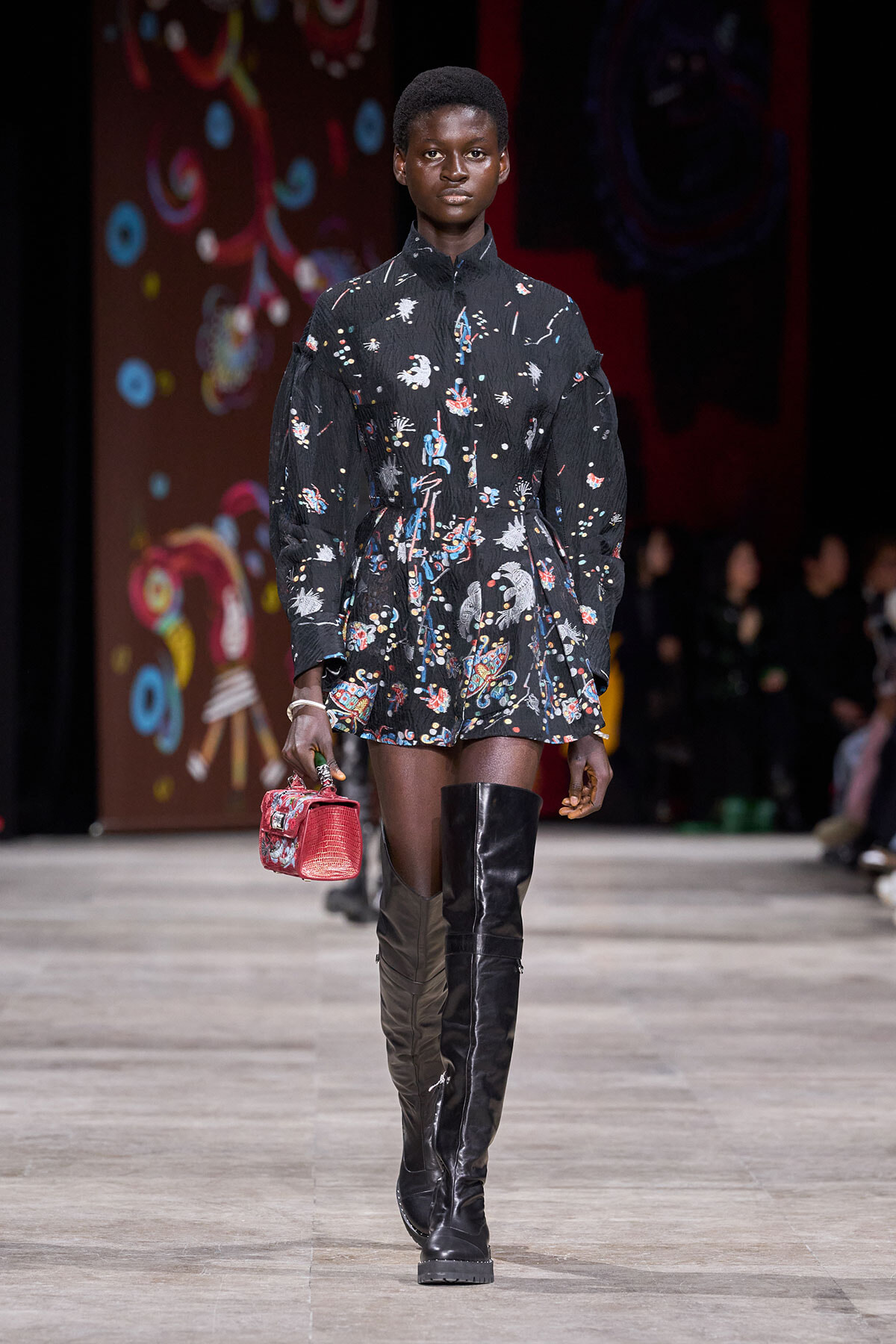 Model walks a runway in a black floral mini dress with voluminous sleeves and over-the-knee black boots, carrying a small red handbag.