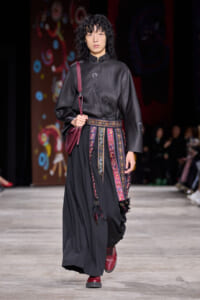 Model walking the runway in a black satin blouse with subtle floral embroidery, paired with a long black skirt and a colorful patterned belt with tassels; burgundy crossbody bag and red boots are visible.