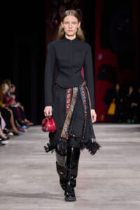 Model walks the runway in a black high-neck jacket and long skirt with a colorful patterned sash, red clutch, and shiny knee-high boots.