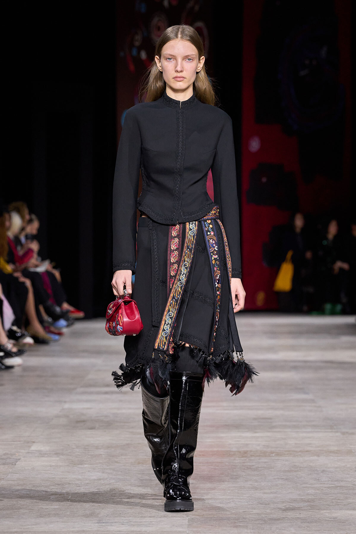 Model walks the runway in a black high-neck jacket and long skirt with a colorful patterned sash, red clutch, and shiny knee-high boots.