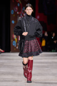 Model walking a runway in a black embroidered bomber jacket, patterned pleated skirt, and tall red knee-high boots with a black crossbody bag.