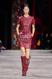 Model walks the runway in a red leather short-sleeve dress with studded trim, paired with red knee-high boots and a small red crossbody bag with chain fringe.