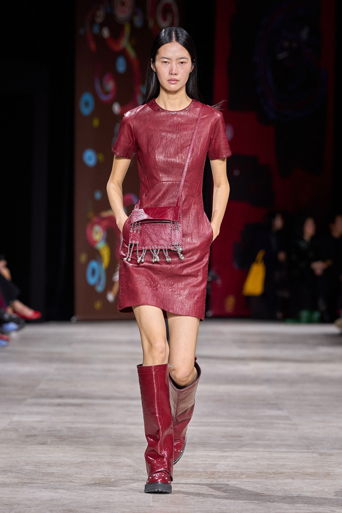 Model walks the runway in a red leather short-sleeve dress with studded trim, paired with red knee-high boots and a small red crossbody bag with chain fringe.
