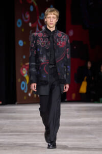 Male model walks the runway in a black embroidered jacket with red patterns, paired with black shirt and trousers.