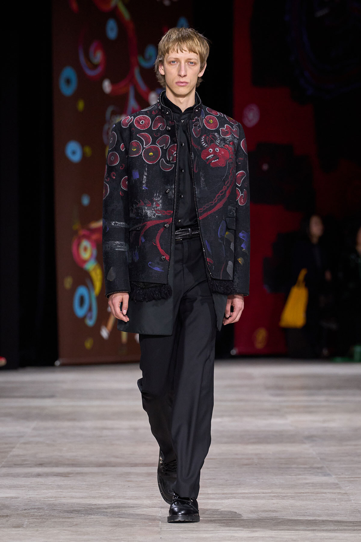 Male model walks the runway in a black embroidered jacket with red patterns, paired with black shirt and trousers.