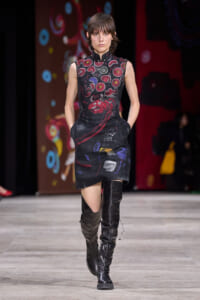 Runway model walks in a sleeveless black dress with red and purple abstract motifs, paired with knee-high black boots.