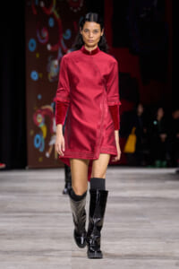 Female model walking a runway in a crimson, mandarin-collar wrap dress with velvet-cuffed sleeves and black knee-high boots with a two-tone finish.