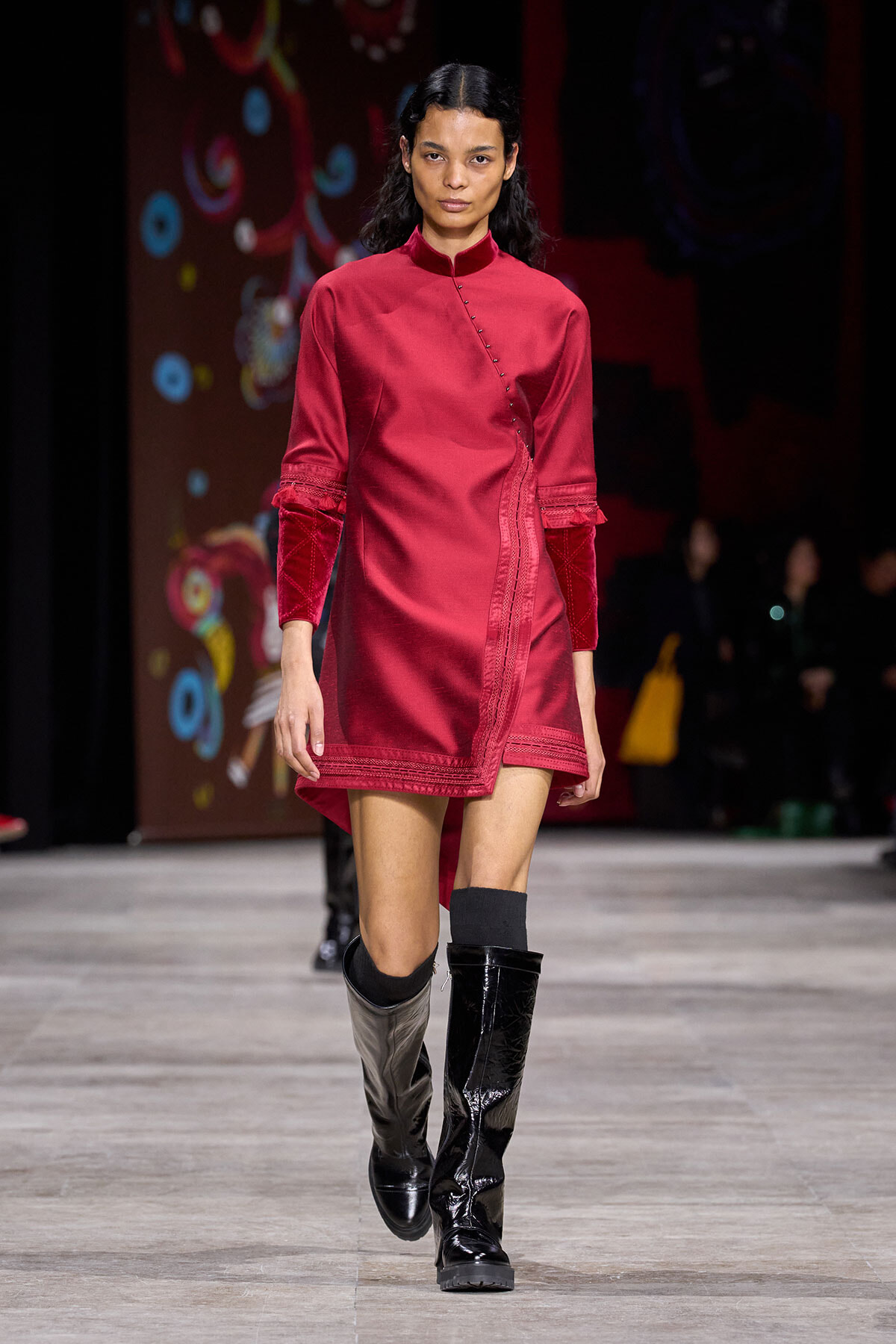 Female model walking a runway in a crimson, mandarin-collar wrap dress with velvet-cuffed sleeves and black knee-high boots with a two-tone finish.