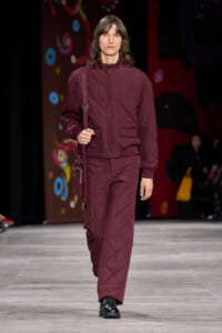 Male model on a fashion runway wearing a maroon quilted jacket and matching pants, carrying a crossbody bag.