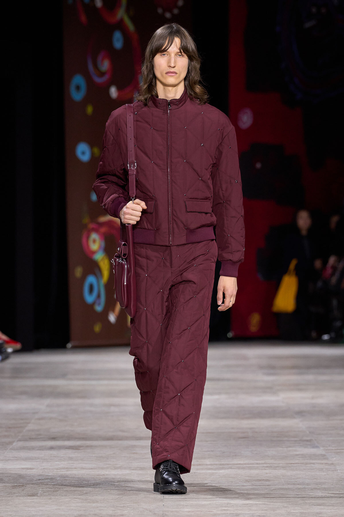 Male model on a fashion runway wearing a maroon quilted jacket and matching pants, carrying a crossbody bag.