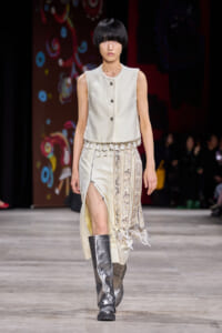 Model walking a runway in a cream sleeveless buttoned top with tassel fringe and a patterned ivory skirt, wearing metallic knee-high boots.