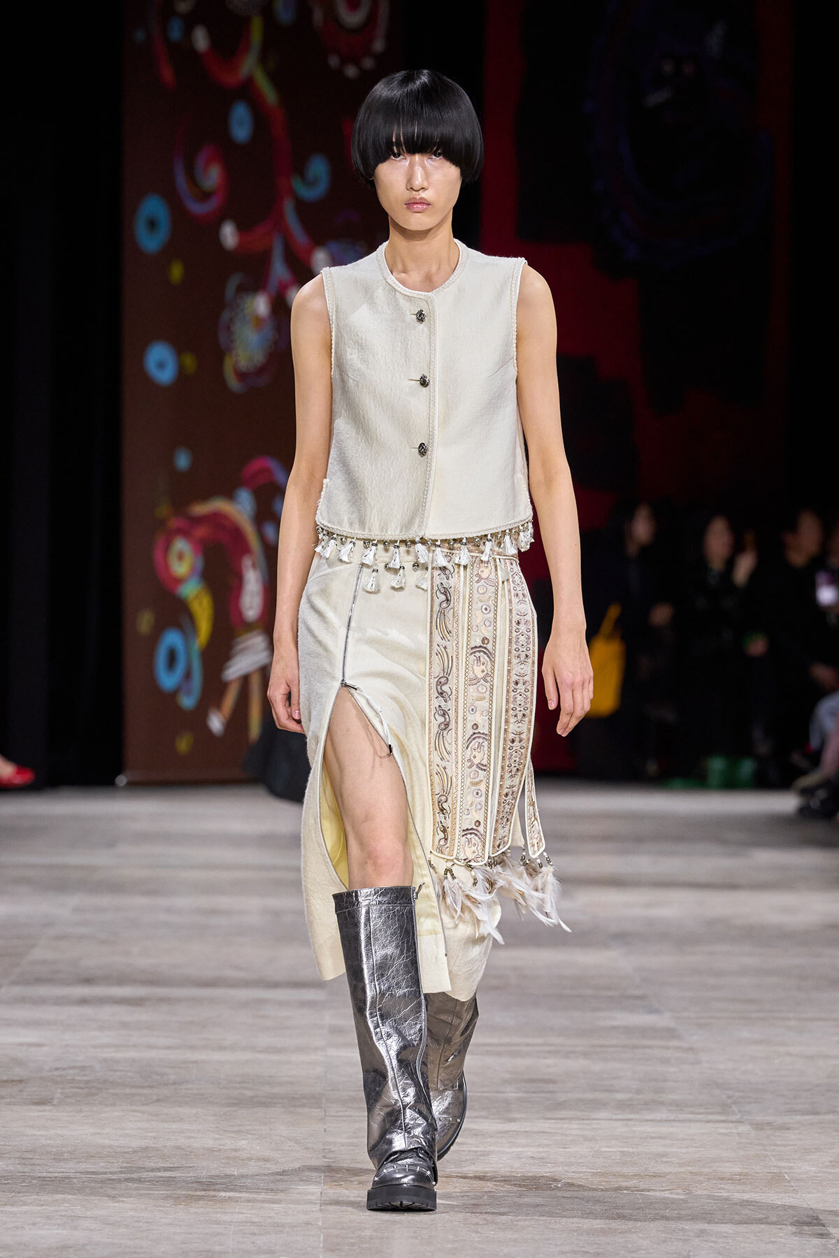 Model walking a runway in a cream sleeveless buttoned top with tassel fringe and a patterned ivory skirt, wearing metallic knee-high boots.