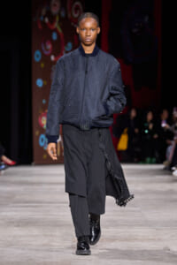 Model walks a fashion runway in a navy quilted bomber jacket with bead details and wide black layered pants, finished with black boots.
