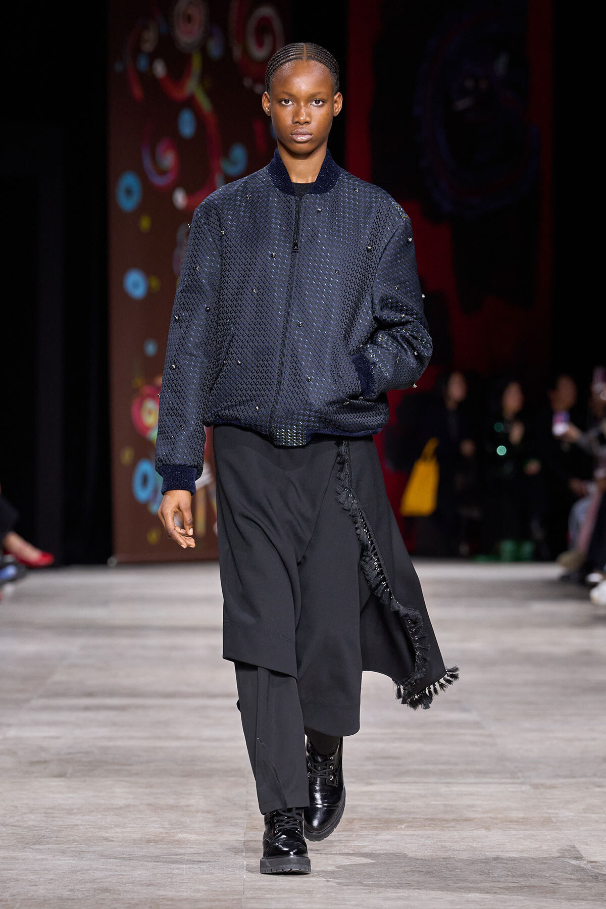 Model walks a fashion runway in a navy quilted bomber jacket with bead details and wide black layered pants, finished with black boots.