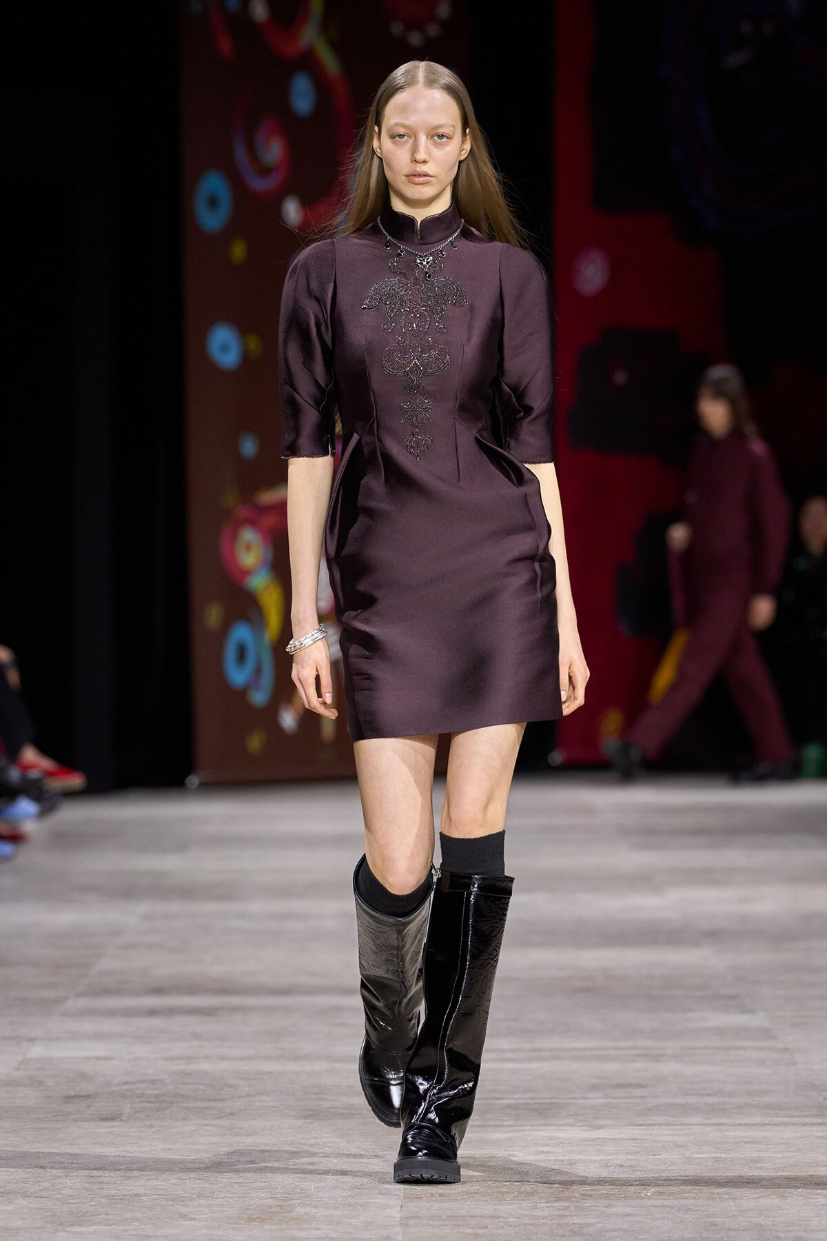 Runway model in a plum satin mini dress with ornate embroidery at the chest, paired with black knee-high patent boots.