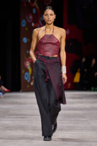 Model walking a runway in a burgundy halter top with beadwork paired with black wide-leg pants and burgundy sash, wearing a chunky silver cuff bracelet.