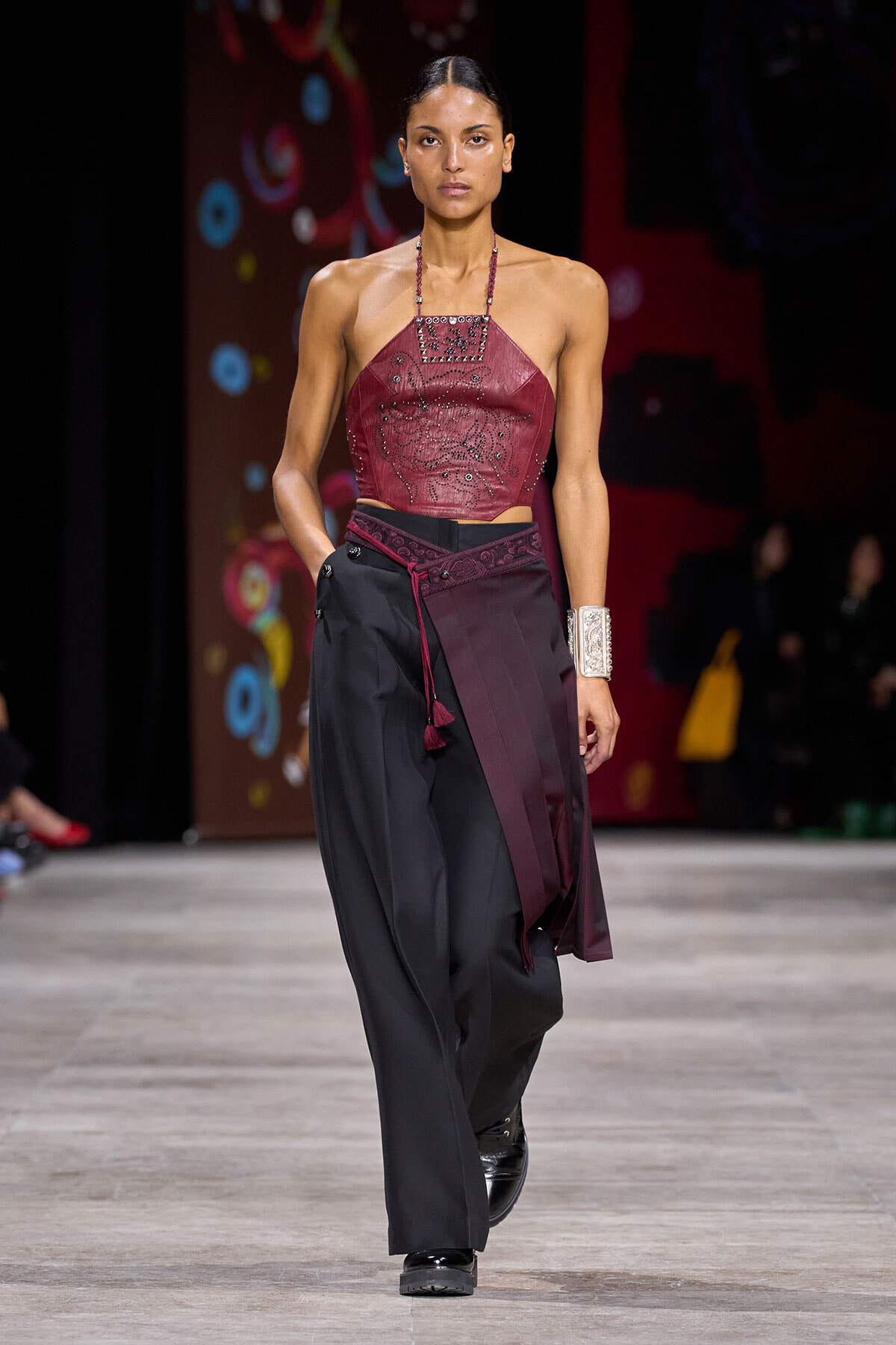 Model walking a runway in a burgundy halter top with beadwork paired with black wide-leg pants and burgundy sash, wearing a chunky silver cuff bracelet.