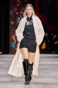 Model on runway wearing a beige long coat over a black shirt and black shorts with a chunky belt and knee-high black boots, posing confidently.