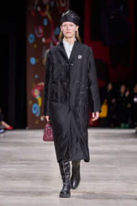 Model on a runway wearing a long black quilted coat with silver embroidery, black cap, white shirt, knee-high boots, and a burgundy handbag.