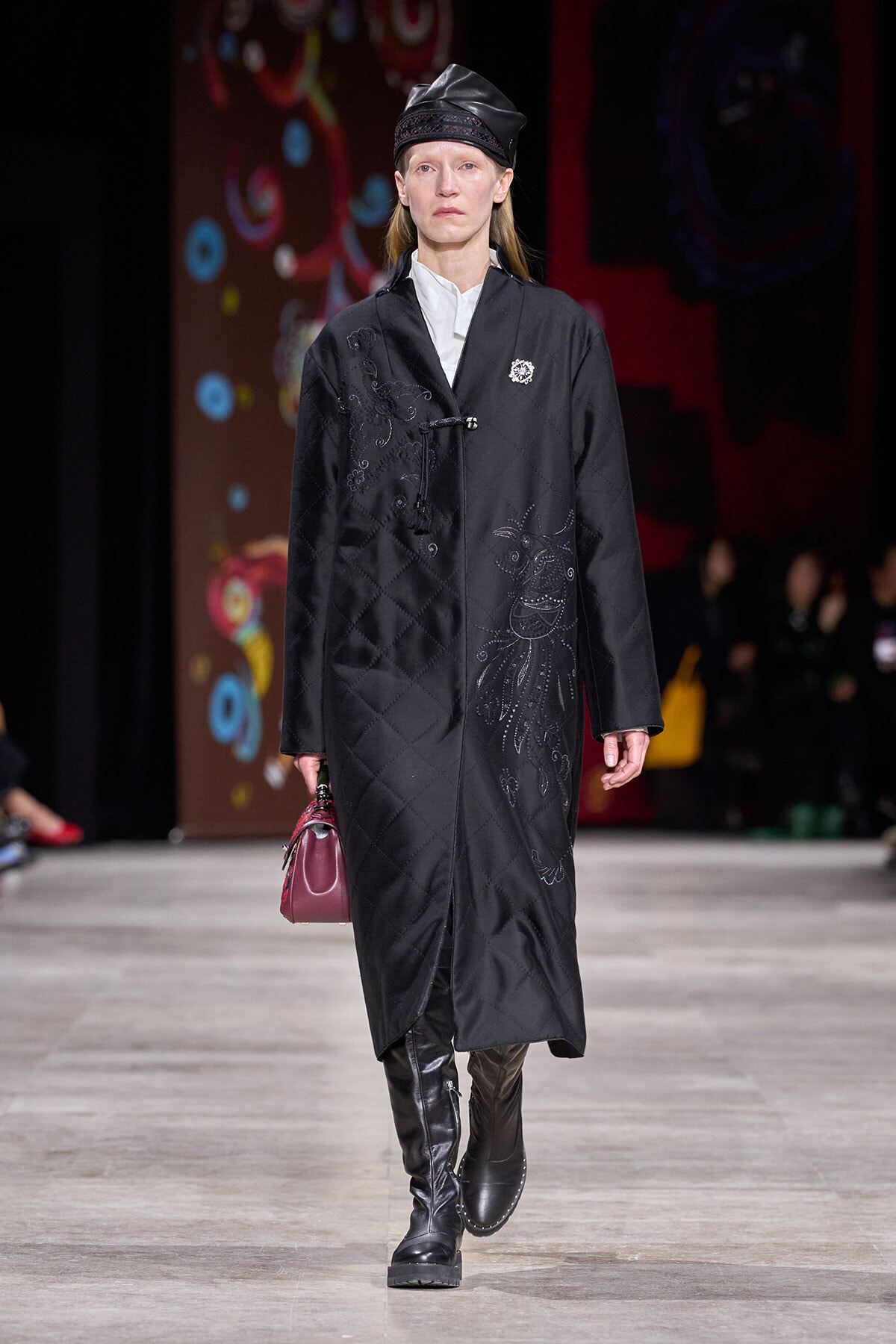Model on a runway wearing a long black quilted coat with silver embroidery, black cap, white shirt, knee-high boots, and a burgundy handbag.