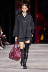 Model walks the runway wearing a dark gray oversized top with matching shorts, black thigh-high boots, and a magenta handbag with chain straps.