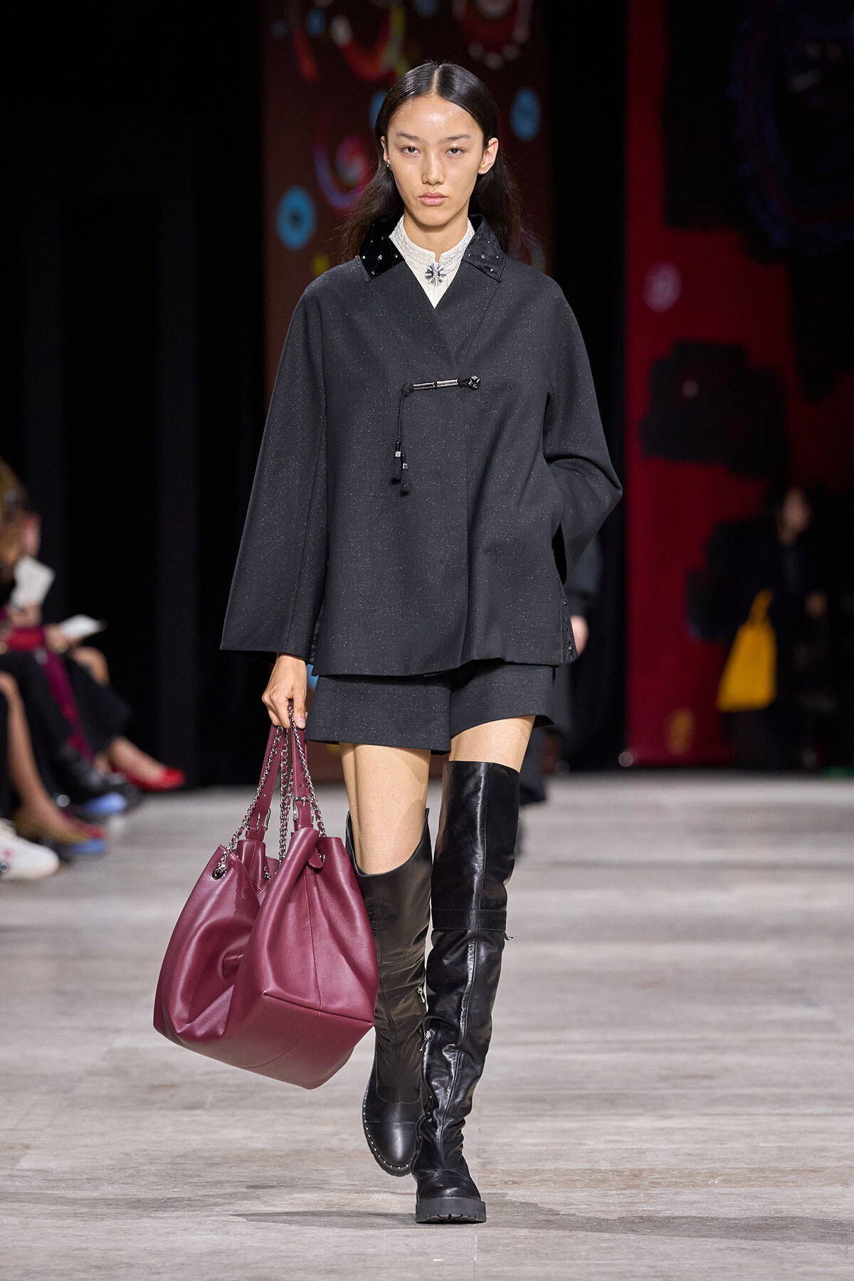Model walks the runway wearing a dark gray oversized top with matching shorts, black thigh-high boots, and a magenta handbag with chain straps.