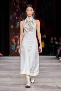 Female model walking a fashion runway in a white high-neck sleeveless gown with an ornate embroidered chest panel and light boots on a dark backdrop runway.