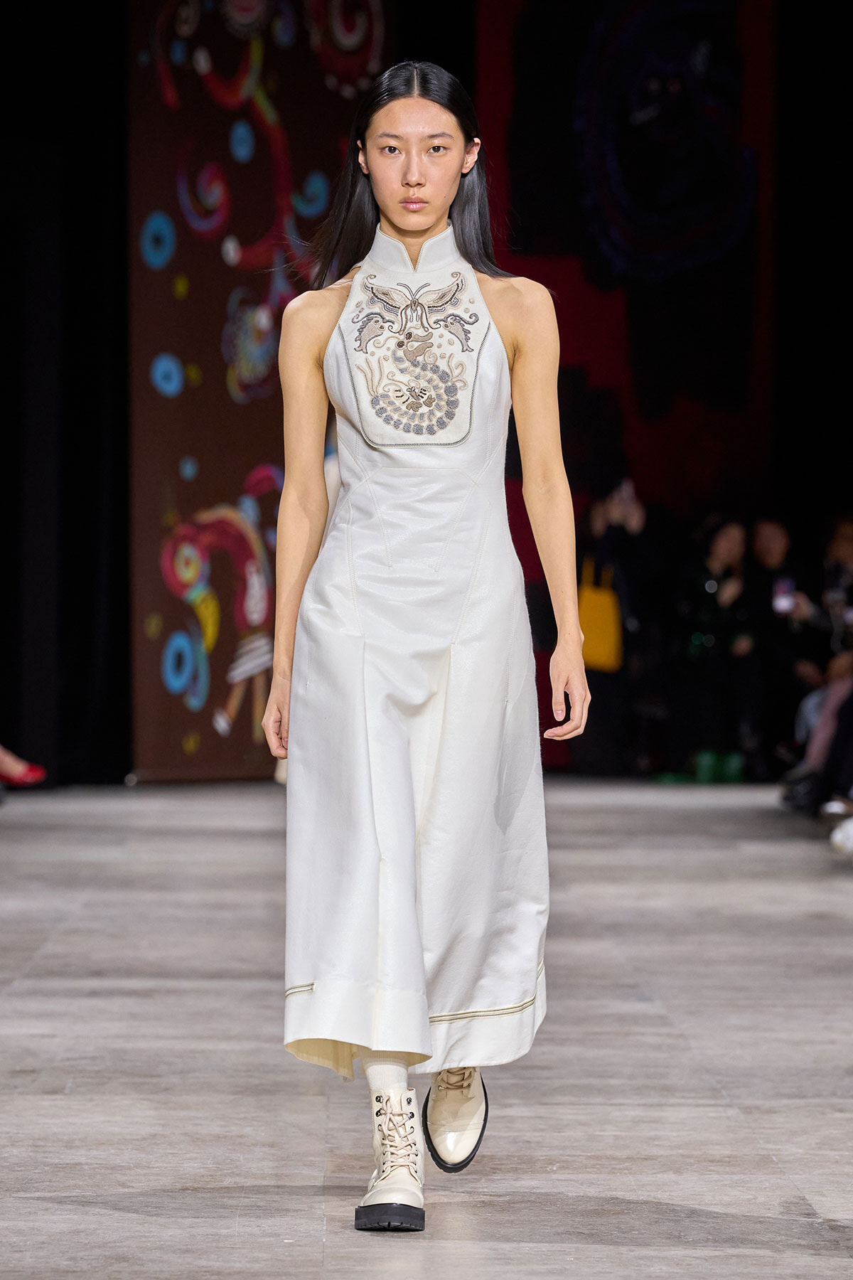 Female model walking a fashion runway in a white high-neck sleeveless gown with an ornate embroidered chest panel and light boots on a dark backdrop runway.