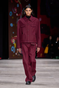 Model walks the runway in a burgundy matching jacket and wide-leg pants with black shoes, neutral expression.