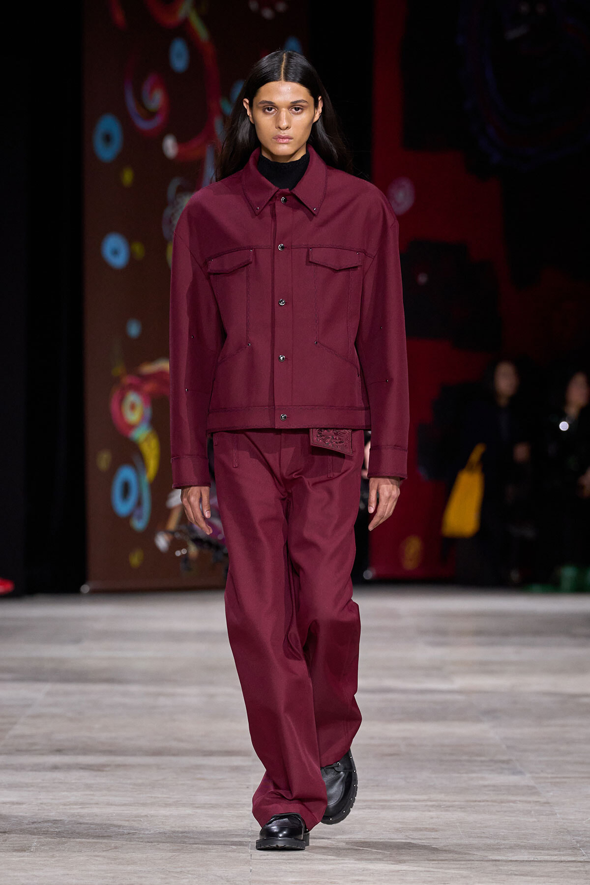 Model walks the runway in a burgundy matching jacket and wide-leg pants with black shoes, neutral expression.