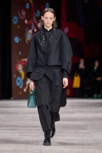 Model in an all-black ensemble with embroidered top, layered skirt, lace-up boots, and a green handbag on the runway.
