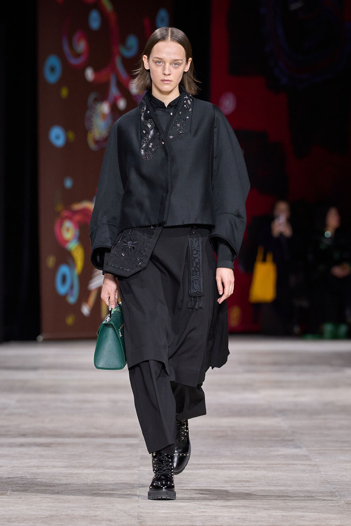 Model in an all-black ensemble with embroidered top, layered skirt, lace-up boots, and a green handbag on the runway.