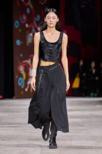 Model walking a fashion show in a black satin crop top with embroidery and a layered black skirt, paired with chunky boots and a silver cuff bracelet on one arm.
