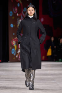 Model on a fashion runway wearing a long black coat with subtle beadwork embroidery and metallic silver boots; hands in pockets as she walks forward.