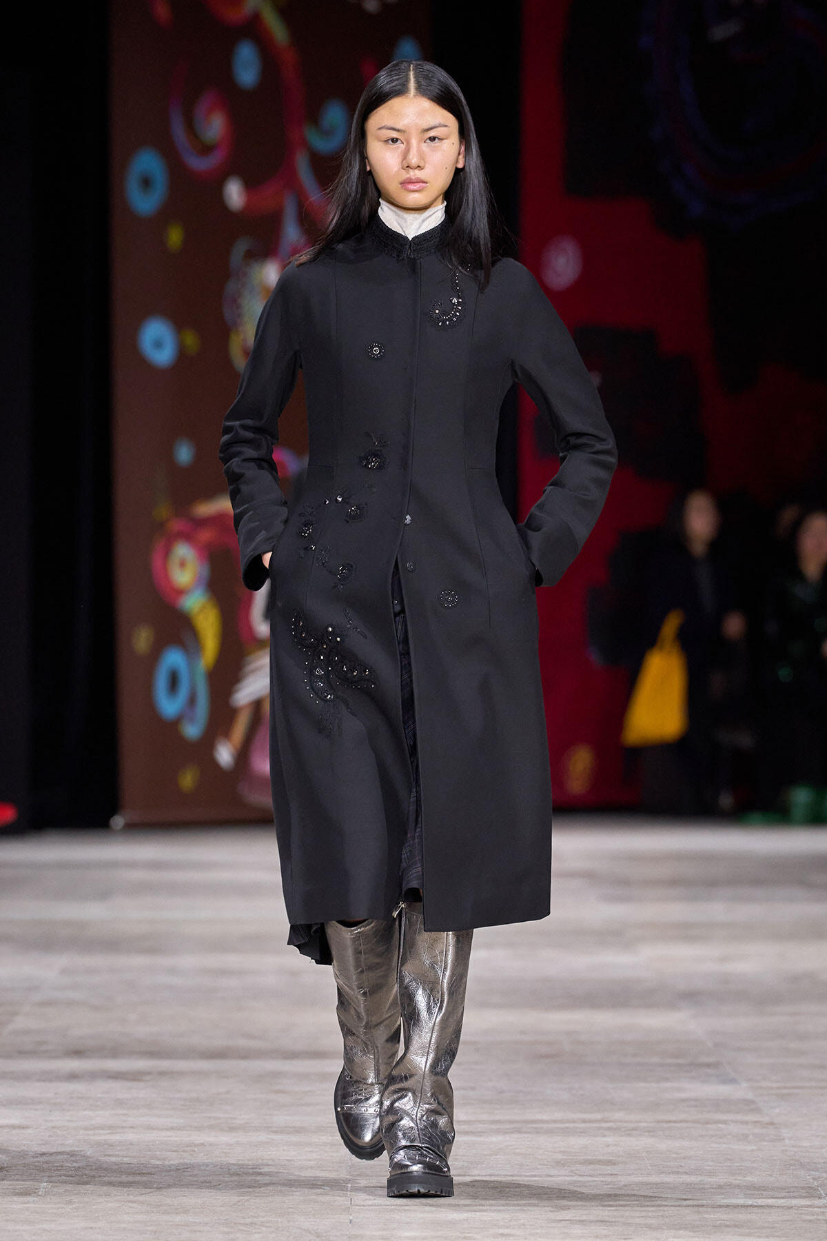 Model on a fashion runway wearing a long black coat with subtle beadwork embroidery and metallic silver boots; hands in pockets as she walks forward.